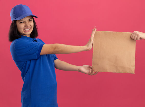 制服身穿蓝色制服 戴着帽子的年轻送货员站在粉色的墙上 拒绝拿一个纸包裹 因为她不满意帽子拿不满意