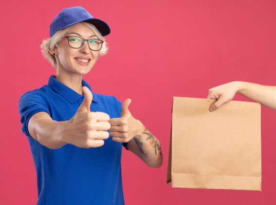 年轻身着蓝色制服 戴着帽子的年轻送货员站在粉红色的墙上 友好地竖起大拇指 一边接受纸袋包装帽子微笑