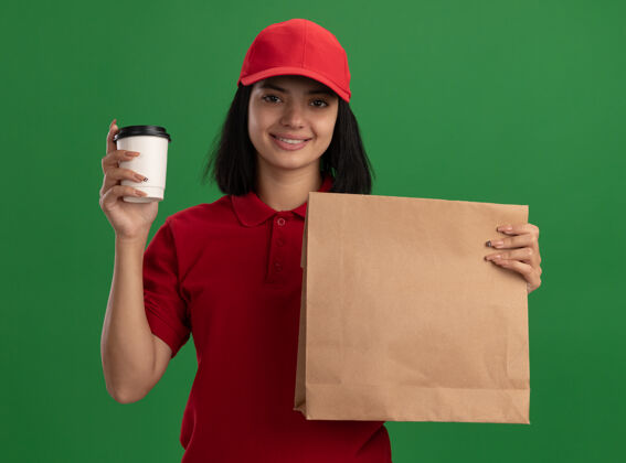 女孩身着红色制服 头戴鸭舌帽 手持纸袋和杯子的快乐少女站在绿色的墙边 欢快地微笑着杯子年轻送货
