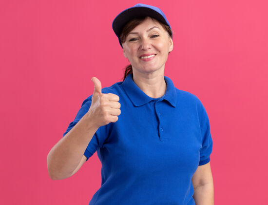 微笑身穿蓝色制服 头戴帽子的中年女送货员站在粉红色的墙上 面带微笑 兴高采烈地竖起大拇指制服站着表演