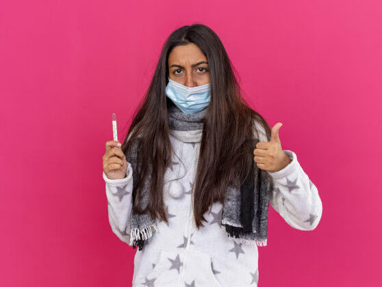 拇指年轻生病的女孩戴着医用面罩 戴着围巾 拿着温度计 在粉色背景上孤立地竖起大拇指 看着摄像机温度计举行围巾