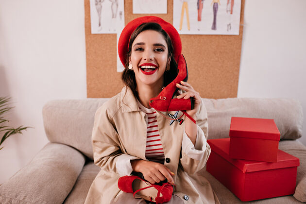 高跟鞋美丽可爱的女士 戴着明亮时尚的贝雷帽 穿着米色秋日战壕 在她的公寓里玩得很开心 还穿着红色的鞋子包成人姿势