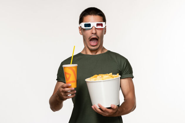 年轻男性正面图身穿绿色t恤的年轻男性手持土豆cips和苏打水 戴着d太阳镜在白色墙上拍摄男性孤独电影电影土豆茶