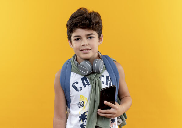 耳机高兴的小男生背着书包 戴着耳机 把电话隔离在黄色的背景上男孩包学校