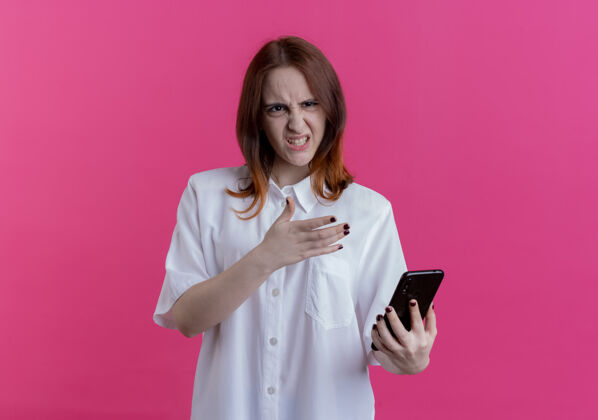 点愤怒的年轻红发女孩拿着手机指着孤立的粉红色背景年轻愤怒红发