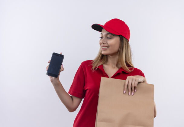 拿着高兴的年轻送货女孩穿着红色制服 戴着帽子 拿着纸袋 看着手机在她的手上孤立的白色背景制服女孩手