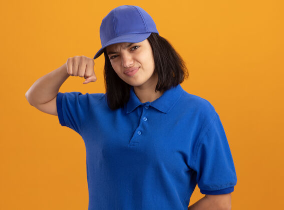 女孩身穿蓝色制服 头戴鸭舌帽 表情严肃自信的年轻送货女孩 站在橘色的墙上挥舞着拳头显示严重交货
