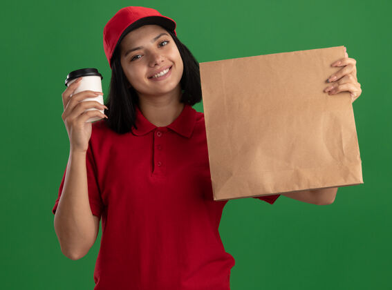 帽子身穿红色制服 头戴鸭舌帽 手持纸袋和纸杯的年轻送货小姐站在绿色的墙边 脸上洋溢着幸福的笑容杯子抱着递送