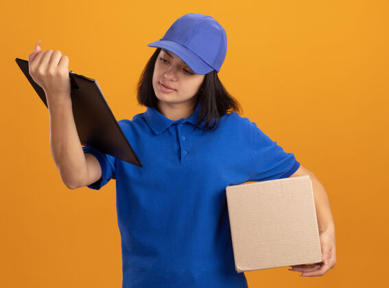 页面穿着蓝色制服 戴着帽子的年轻送货女孩拿着纸板箱和空白页的剪贴板 严肃地看着站在橙色墙上的页面空白帽子交货