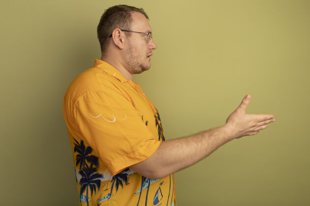 戴一个戴着眼镜 身穿橘色衬衫的男人站在一边 举起手 越过绿色的墙问手眼镜男人