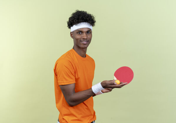 穿高兴的年轻黑人美国运动男子戴着头带和手环举行乒乓球拍与球隔离在绿色背景请腕带乒乓球
