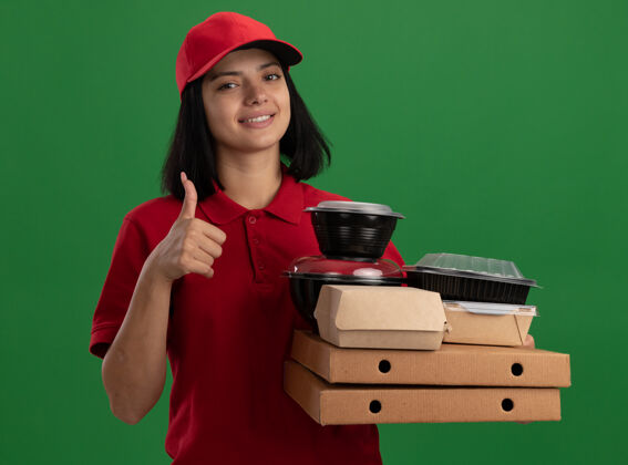 盒子身穿红色制服 头戴鸭舌帽的年轻送货员 手拿披萨盒和食品包装 脸上带着微笑 竖起大拇指站在绿色的墙上送货站着微笑