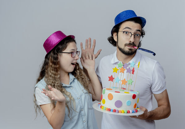 年轻人快乐的年轻夫妇戴着粉色和蓝色的帽子男孩给女孩生日蛋糕和吹哨子隔离在白色背景上蛋糕欢乐女孩