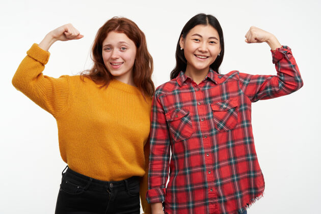 快乐两个年轻快乐的女性朋友穿着黄色毛衣和格子衬衫展示她们有多强壮 肌肉发达充满活力人的概念隔着白墙孤立毛衣活力年轻