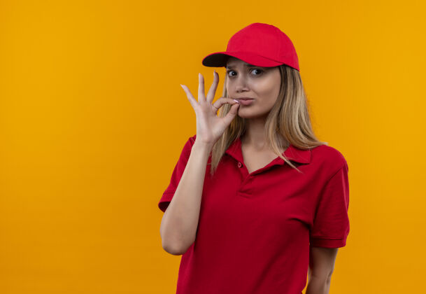 送货看着镜头 穿着红色制服 戴着帽子的年轻送货女孩在橙色背景上显示出一种很好的姿势秀帽子制服