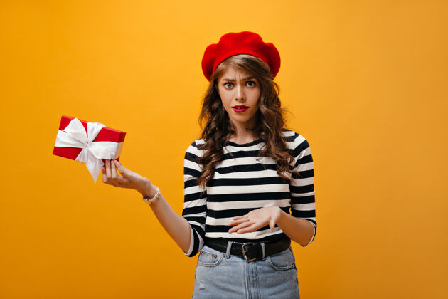 女性女孩的肖像看起来很不开心 拿着礼盒穿着红色贝雷帽和牛仔裙 带着黑色腰带的现代年轻女子摆姿势表情礼物帽子