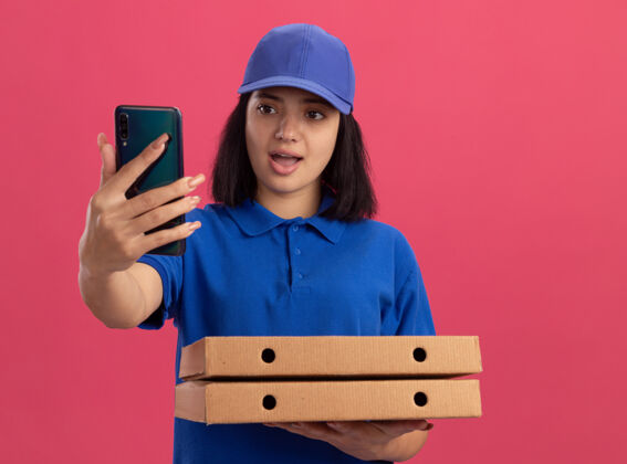 站着身穿蓝色制服 头戴鸭舌帽 拿着比萨饼盒的年轻送货员站在粉色的墙上 困惑地看着智能手机的屏幕送货拿着帽子