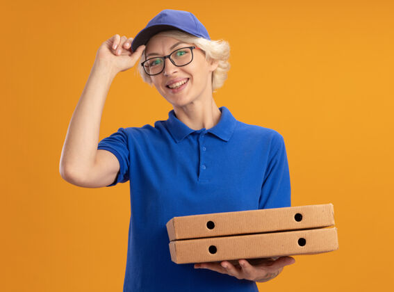 自信身穿蓝色制服 戴着眼镜 拿着比萨饼盒的年轻送货员自信地微笑着 把帽子戴在橙色的墙上盒子送货拿着