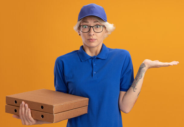 披萨身穿蓝色制服 戴着眼镜 拿着比萨饼盒的年轻送货女被人和举过橙色墙壁的手臂搞混了送货胳膊帽子
