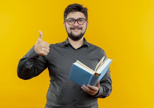 年轻人高兴的年轻商人戴着眼镜拿着书大拇指孤立地站在黄色的背景上眼镜戴着拇指