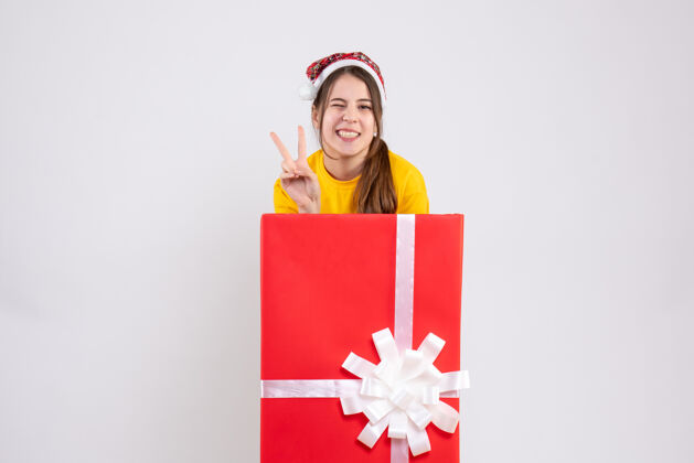 礼物前视图幸福的女孩与圣诞帽制作胜利标志站在背后大圣诞礼物惊喜胜利铅笔