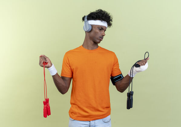 手臂困惑的年轻黑人运动男子戴着头带和腕带和电话臂带与耳机举行 看跳绳隔离在绿色背景腕带头带举行