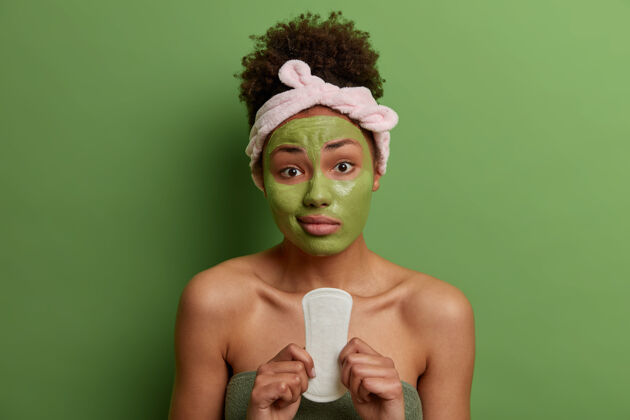 身体不高兴的年轻女子拿着月经卫生巾 敷上美容面膜 戴上头带和毛巾 在室内对着绿墙摆姿势女人 美丽 卫生理念棉花成人年轻