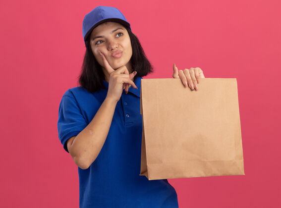 拼图穿着蓝色制服 戴着帽子 拿着纸包裹的年轻送货女孩站在粉红色的墙上 困惑地看着一旁制服帽子年轻