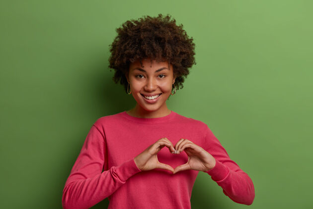 肖像美丽的非洲裔美国女人的镜头表达爱 在浪漫的心情 显示心的迹象 坦白的真实感情 有同情心 穿着玫瑰色的套头衫 对绿墙姿势非洲高兴浪漫