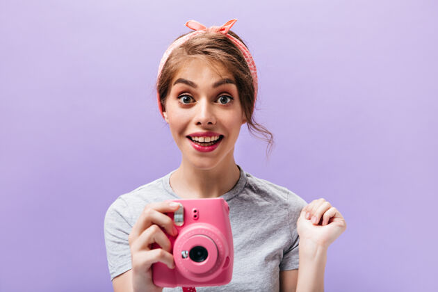 衬衫穿着灰色衬衫的迷人女人手持粉色相机戴着夏日头巾的快乐漂亮女孩穿着时髦的衣服摆姿势与世隔绝的背景成人脸相机