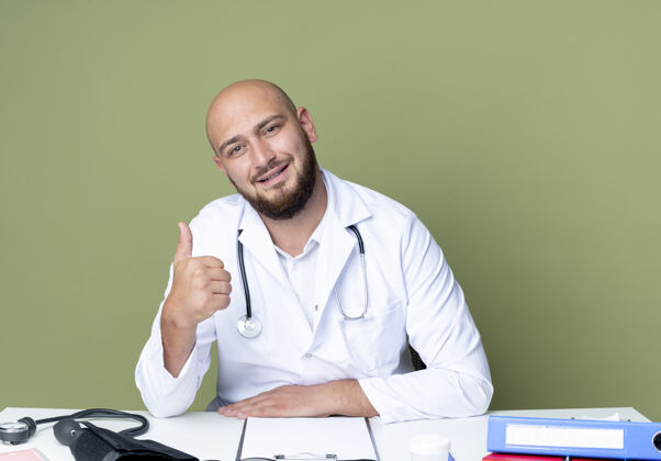 拇指高兴的年轻秃头男医生穿着医用长袍和听诊器坐在办公桌前用医用工具工作 他的大拇指孤立地竖起在绿色的背景上工具医生秃头