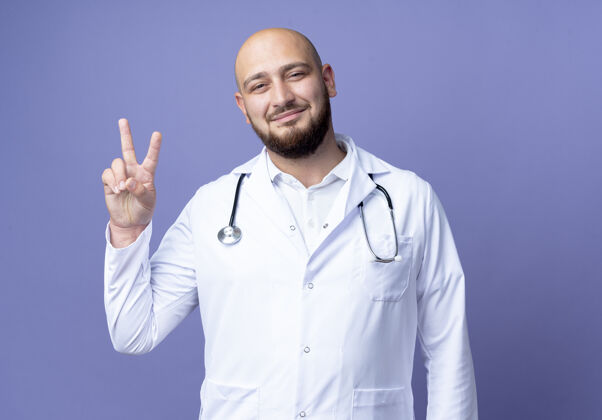 年轻高兴的年轻秃头男医生穿着医用长袍和听诊器显示出奥基手势隔离在蓝色背景男性医疗医生