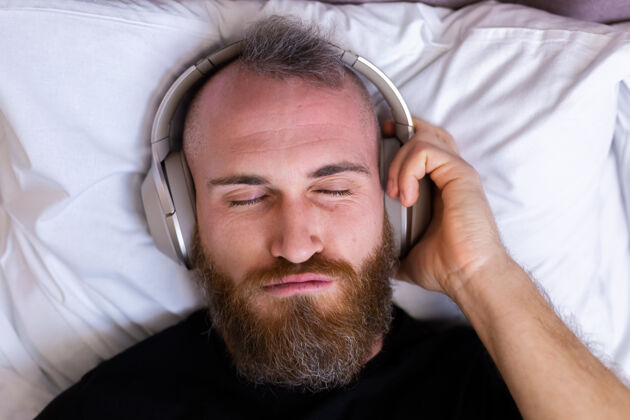 舒适快乐的白人男子戴着耳机躺在床上听他最喜欢的音乐 独自休息 跳舞房子移动无线