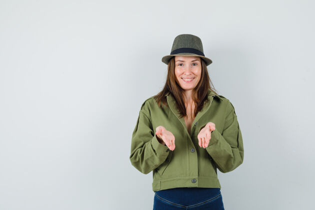 帽子穿着夹克 裤子 帽子假装提供或展示东西的年轻女士 看上去很高兴积极一起夹克