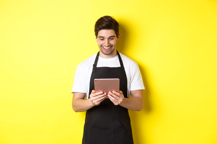 模特穿着黑色围裙的卖家看着数码平板电脑屏幕 微笑着高兴 站在黄色背景上工作室成人帅哥