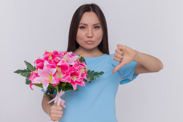 国际一位身穿蓝色连衣裙的亚裔妇女手持花束 不高兴地朝前伸大拇指 站在白色的墙上庆祝国际妇女节站立向下花束