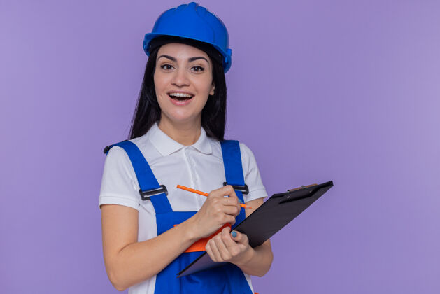微笑身穿建筑制服 头戴安全帽 拿着写字板和铅笔的年轻建筑妇女站在紫色的墙上 面带微笑地看着前方女人建筑工欢呼