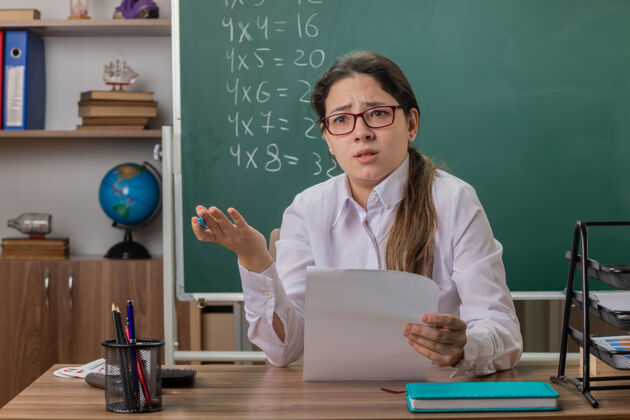 眼镜戴着眼镜的年轻女教师坐在教室的黑板前 一副茫然而不高兴的样子空白前面书桌