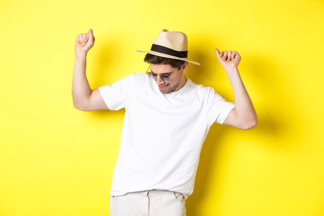 工作室旅游 旅游和度假概念男人旅游者享受假期 戴着草帽和太阳镜跳舞 在黄色背景下摆姿势模特太阳镜夏天
