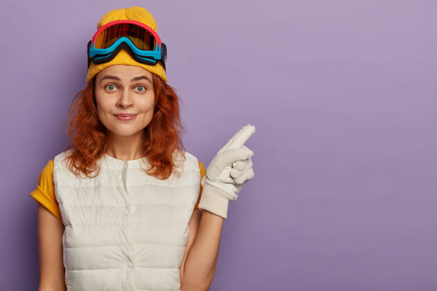 手套迷人的红发女人戴着暖和的帽子 滑雪镜和白色套头衫肖像指向成人