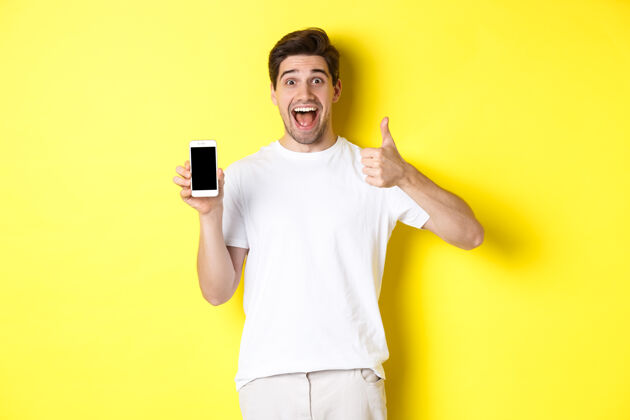 竖起大拇指快乐的年轻人展示拇指和手机屏幕 推荐应用程序或互联网站立男人帅气