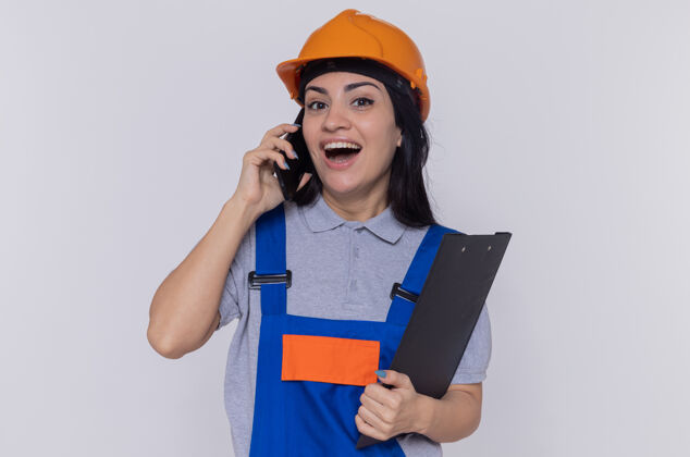 建筑工人身穿施工制服 头戴安全帽的年轻建筑工人站在白色的墙壁上 手里拿着写字板 一边高高兴兴地说着手机电话手机制服