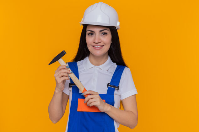 制服身穿施工制服 头戴安全帽 手持铁锤 面带微笑地站在橙色墙壁上的年轻建筑工人头盔锤子建筑工人