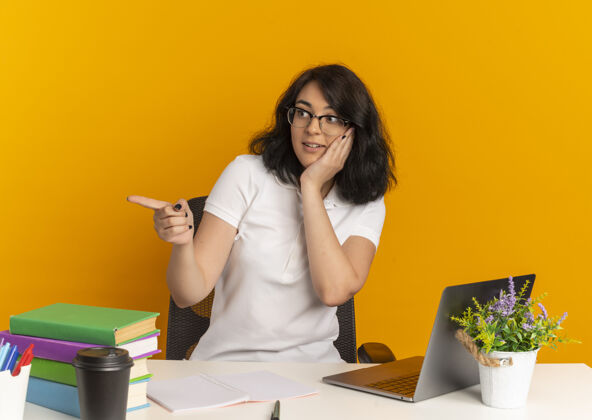 桌子年轻漂亮的白人女生戴着眼镜坐在书桌旁 手里拿着学习工具 指着脸 看着橙色的一面和复印空间复制惊喜工具