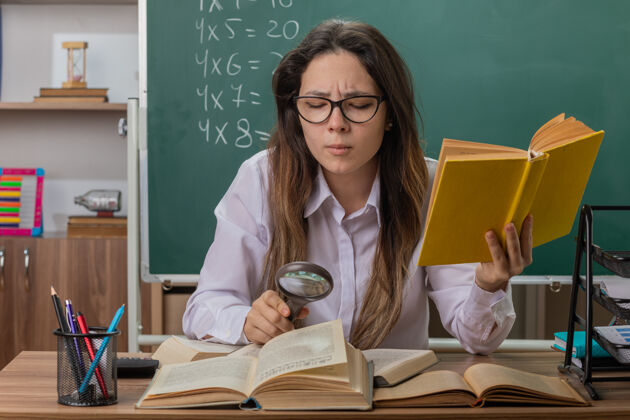 放大镜年轻的女老师戴着眼镜 坐在教室黑板前的课桌旁 透过放大镜看书 很感兴趣穿黑板学校