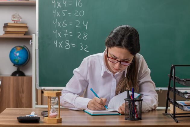 写作戴眼镜的年轻女老师在备课笔记本上写东西 坐在教室黑板前的课桌旁 看起来很自信书桌学校穿