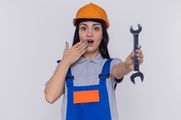 女人年轻的建筑工人穿着建筑制服 戴着安全帽 拿着扳手看着前面 惊讶地捂住嘴 手站在白色的墙上拿着安全嘴