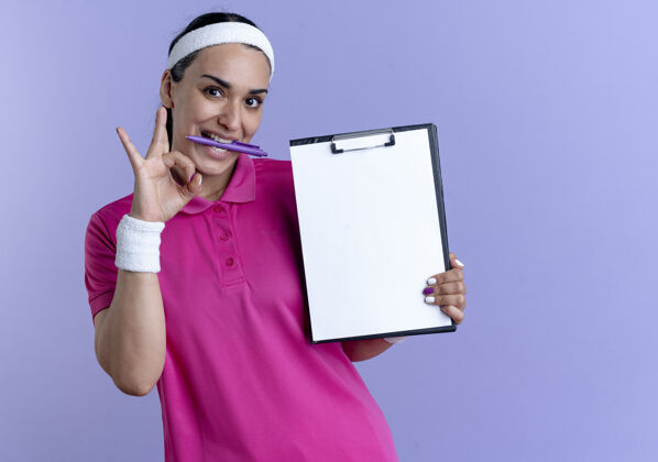 剪贴板年轻快乐的白人运动女性戴着头带和腕带拿着钢笔用牙齿做手势ok手语拿着剪贴板在紫色上有复印空间运动欢乐钢笔