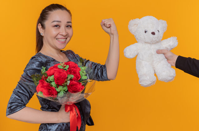 站立快乐和兴奋的亚洲妇女期待收到泰迪熊作为礼物微笑着高兴地握紧拳头庆祝国际妇女节站在橙色的墙上拳头欢呼亚洲