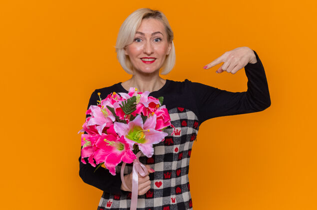 庆祝身着漂亮衣服的快乐年轻女子手持花束 用食指指着花束 站在橘红色的墙上 欢快地笑着庆祝国际妇女节举行衣服欢呼